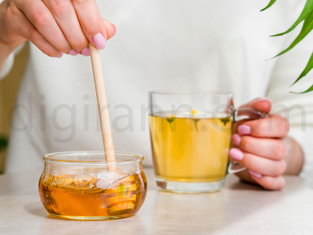نمایی از خوردن ترکیب عسل و آب گرم با قاشق چوبی و ظرف شیشه ای عسل و لیوان ماگ آب توسط زن با لباس سفید و لاک صورتی