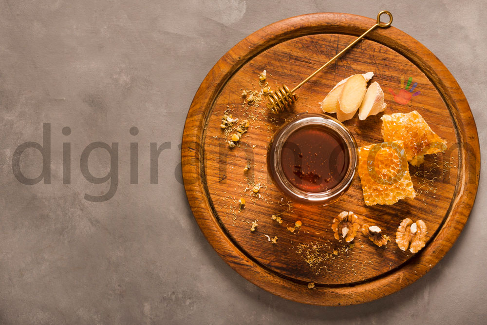 نمایی از خواص عسل کنار در سینی چوبی دایره ای با گردو و تکه های موم عسل و قاشق عسل طلایی