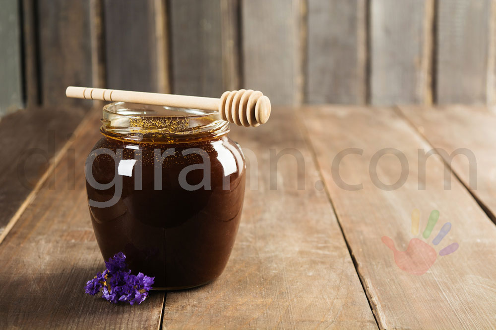 نمایی از ظرف یک کیلوگرمی عسل سیاه روی میز چوبی با قاشق چوبی عسل روی آن